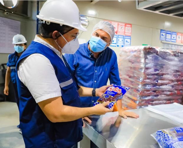 Two team members in hard hats, hairnets and face masks inspect a bag of beans at the facility, next to pallets of product.